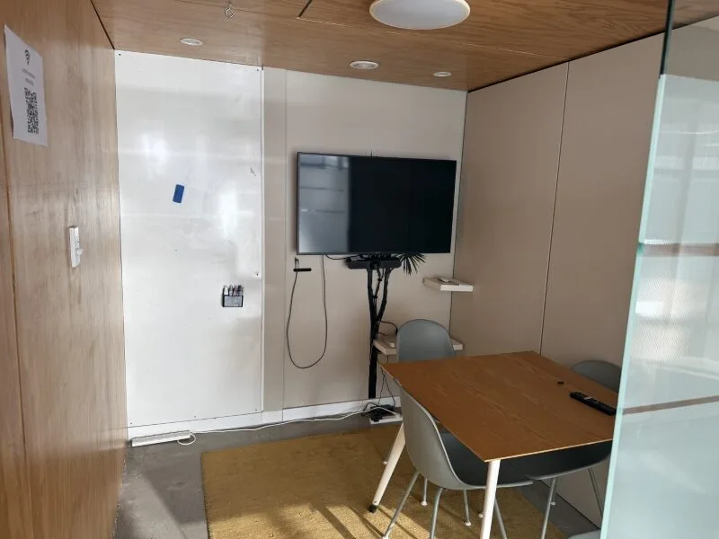Private meeting room at Lyall Bay Work Hub in Wellington with wall-mounted TV screen, whiteboard wall, table seating for four, and modern glass partition for collaborative sessions.